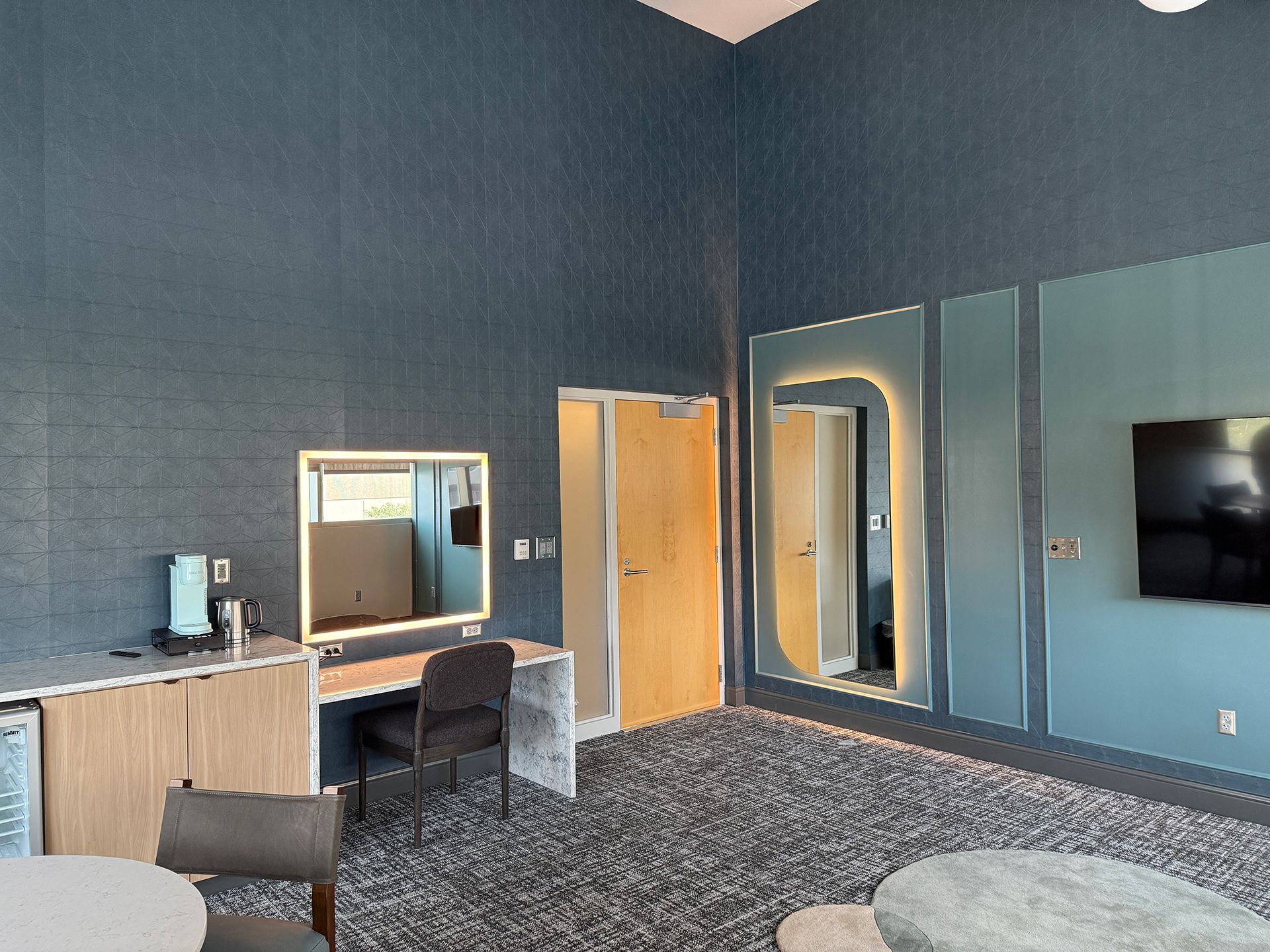 Blue-walled room with a desk, chair, mirror, and TV, with dark carpet and wood doors.