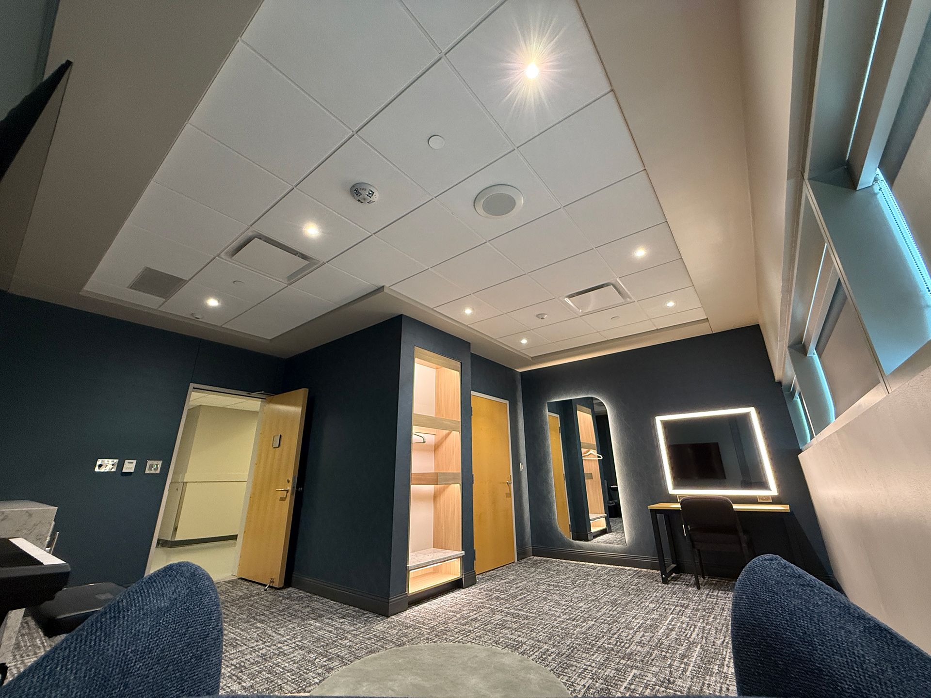 Modern lounge with dark walls, patterned carpet, ceiling lights, built-in shelves, and a lit vanity mirror.