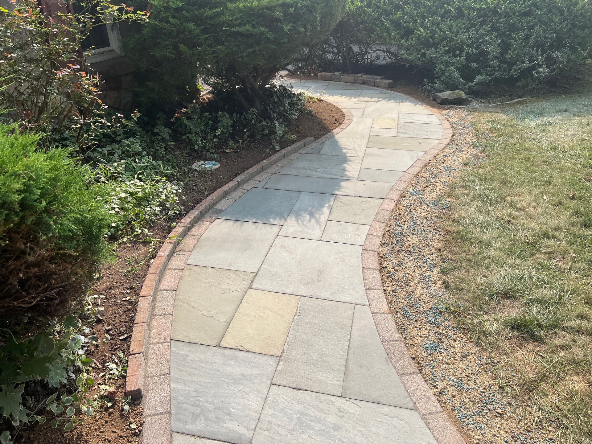 Stone pathway leads to a house entrance, flanked by flowerbeds with various green and yellow plants, and black mulch.