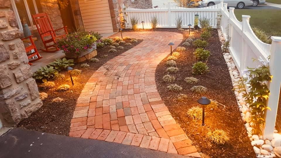 Brick pathway leading to a house, lit with garden lights, flanked by flower beds and white picket fence.