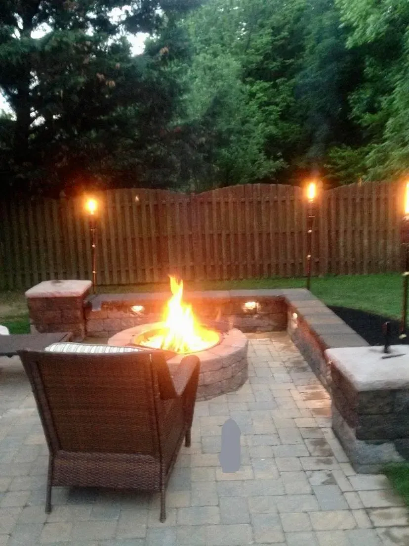 Backyard patio with fire pit, seating, stone pavers, and tiki torches. Green trees and a wooden fence.