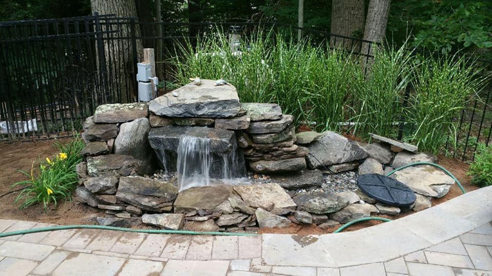 Stone waterfall feature in a backyard, flowing water, surrounded by rocks and plants, near a paved patio.