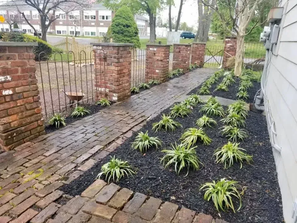 Brick pathway lined with flower beds, leading to a metal gate and brick pillars.