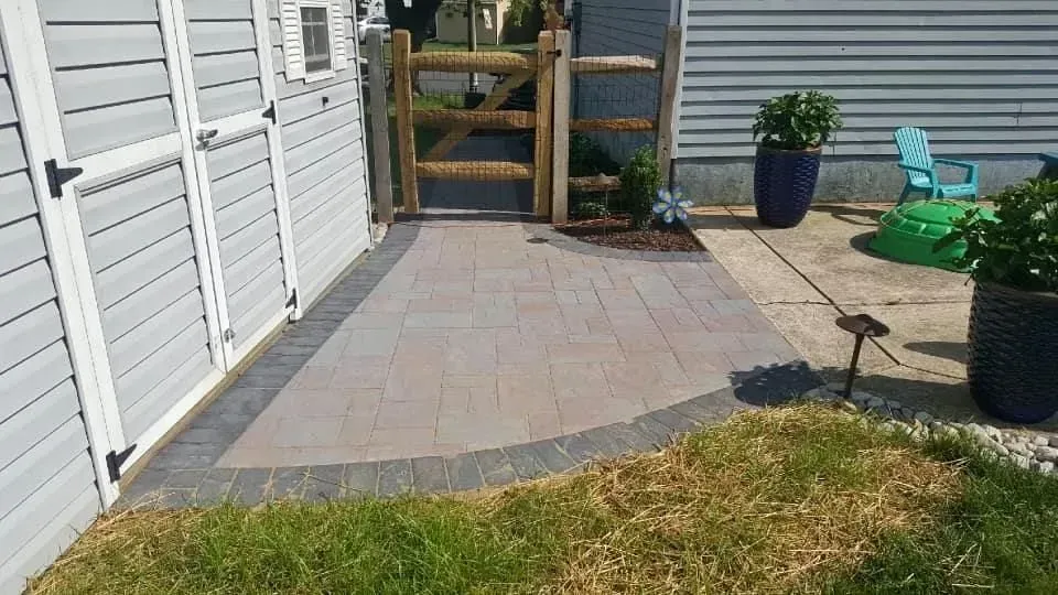 Paver patio with curved edge and wooden gate. Side of a house with two large planters.