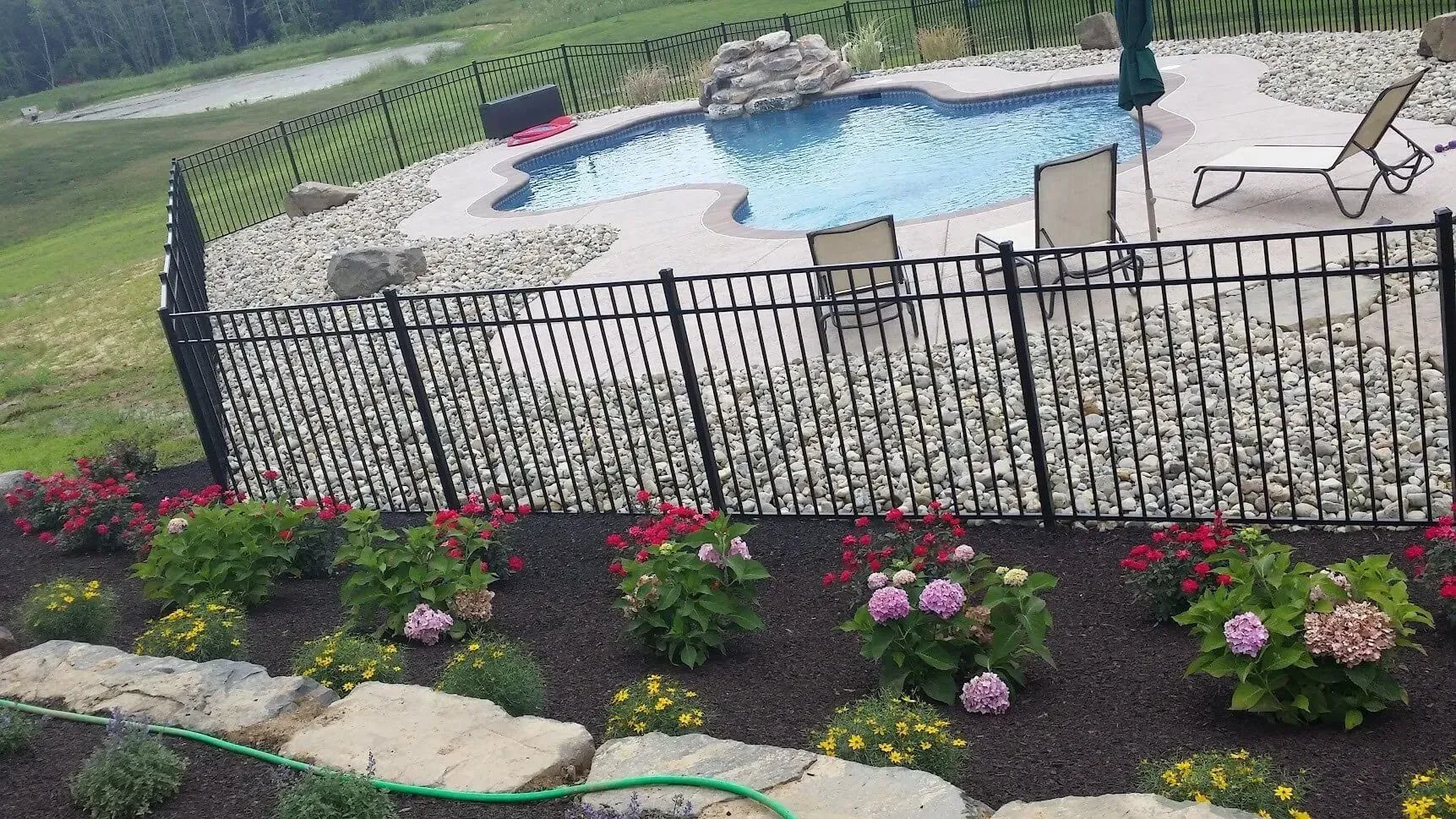 Pool with rock surround, black fence, and colorful flowerbeds on a hillside.