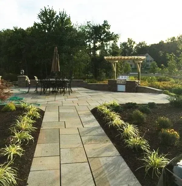 Stone patio with a dining set, built-in grill, pergola, and landscaped garden.