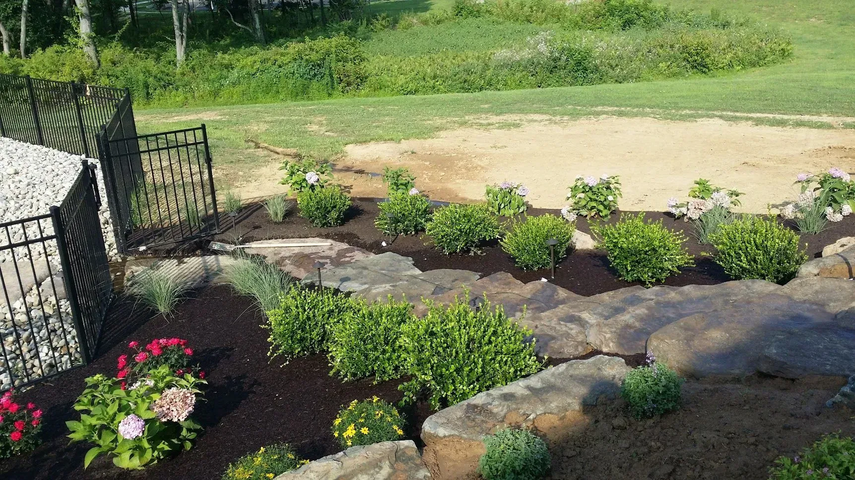 A landscaped garden bed with dark mulch, green shrubs, and stone edging, next to a black fence.