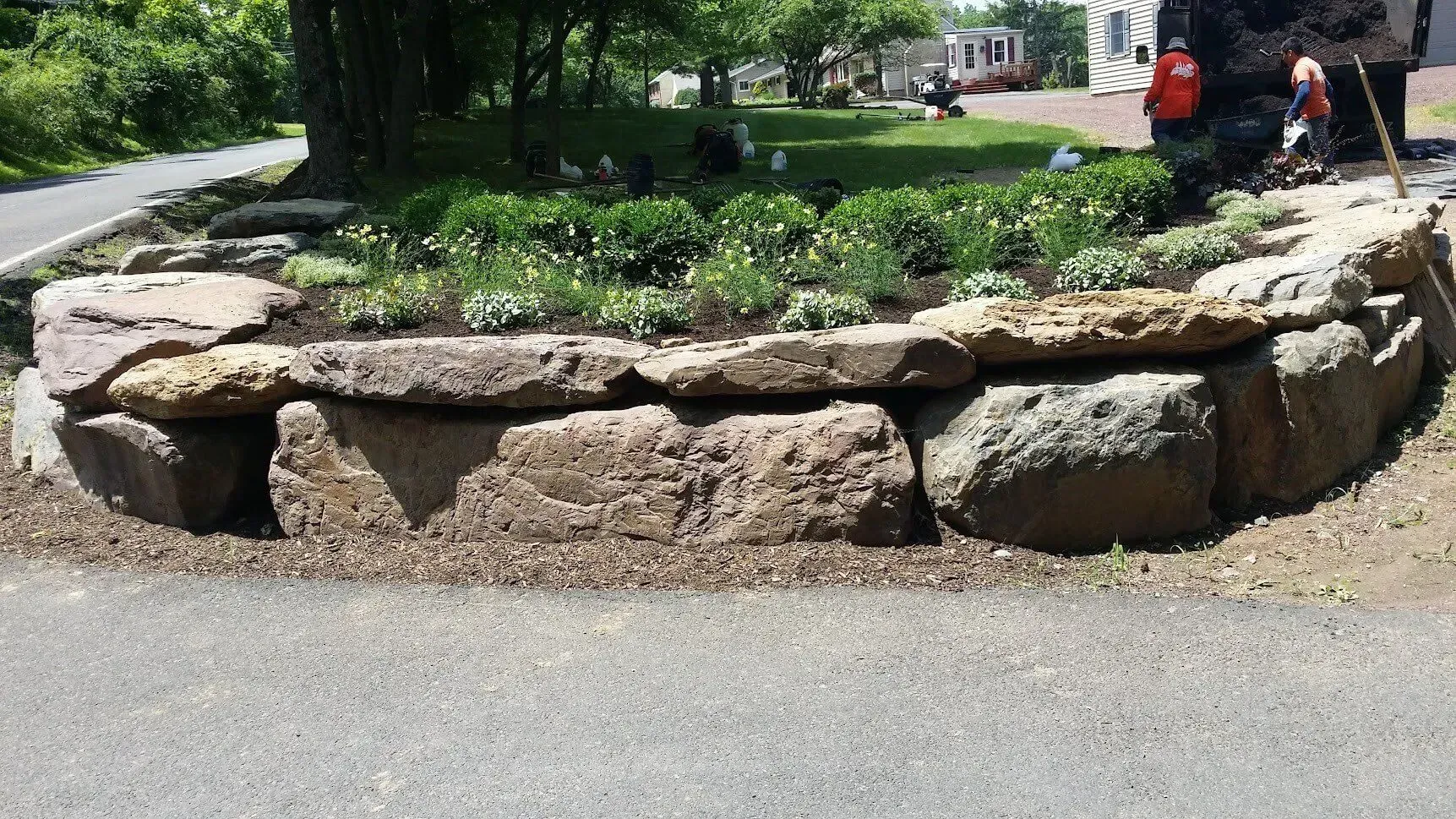 Stone retaining wall bordering a garden bed with green plants, asphalt path in foreground.