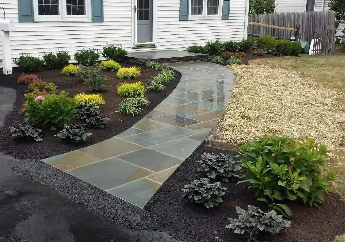 Stone pathway leads to a house entrance, flanked by flowerbeds with various green and yellow plants, and black mulch.