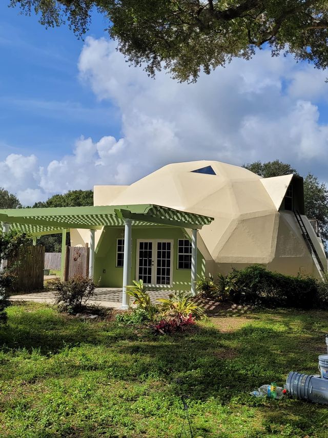 Tan geodesic dome home with a green porch, set in a yard with grass and trees under a blue sky.