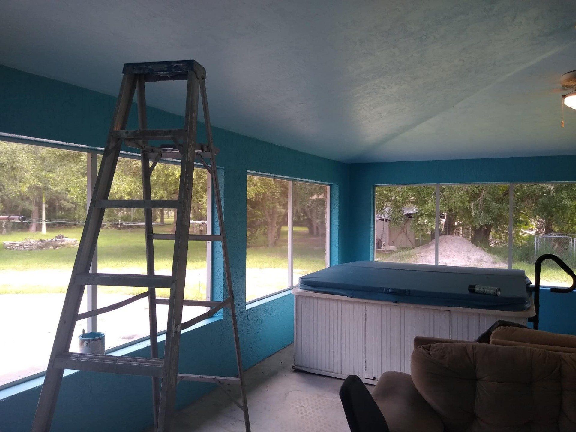 A teal-painted sunroom with a ladder, hot tub, and large windows overlooking a backyard.