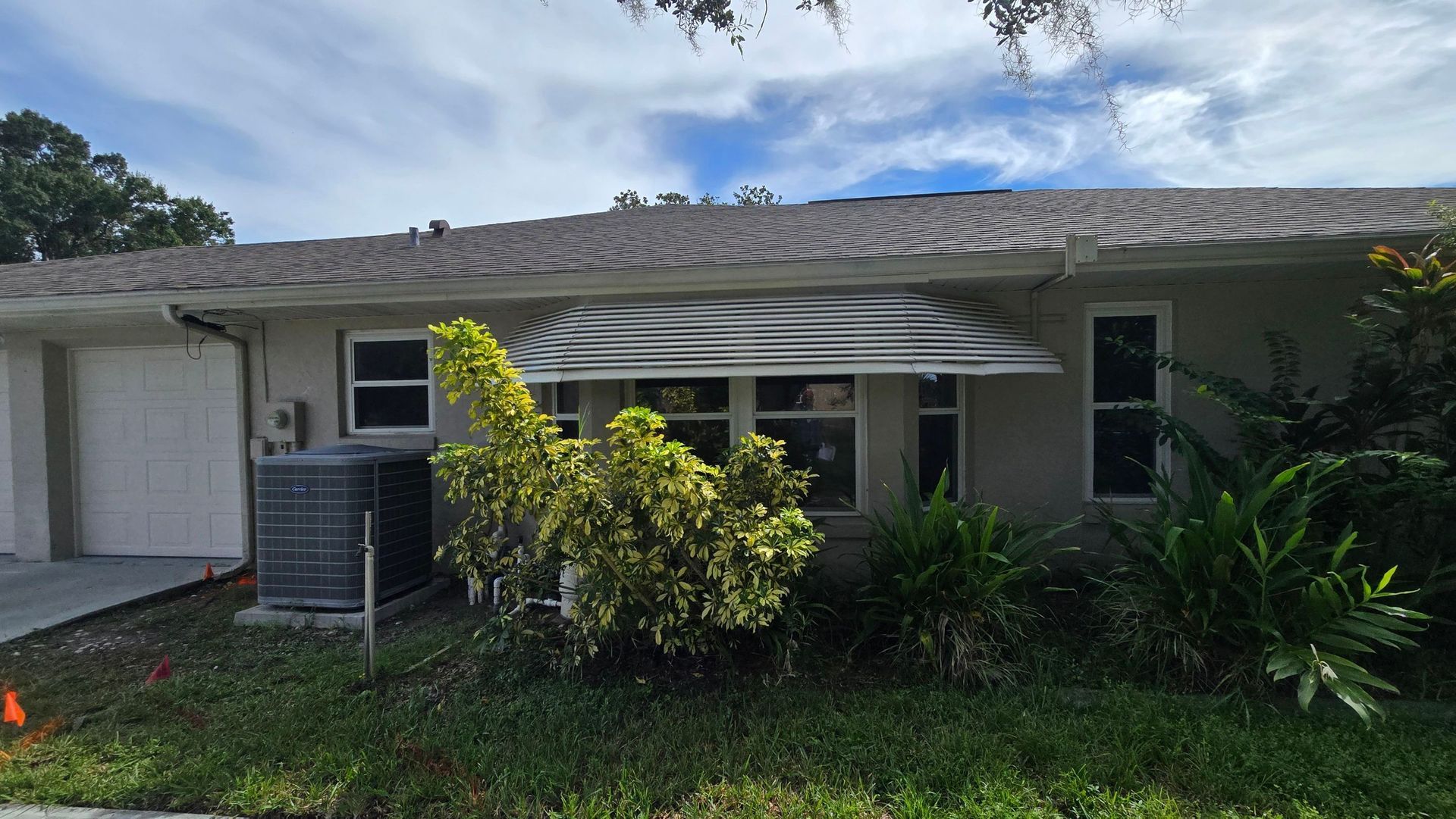 A beige house with a clear awning over windows, green bushes, and a gray air conditioning unit.