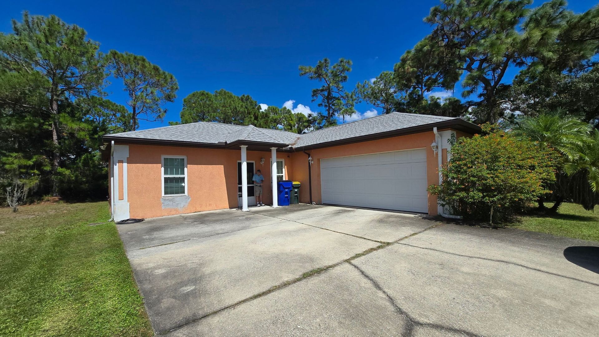 Orange stucco house with gray roof, white garage door, and driveway, surrounded by trees.