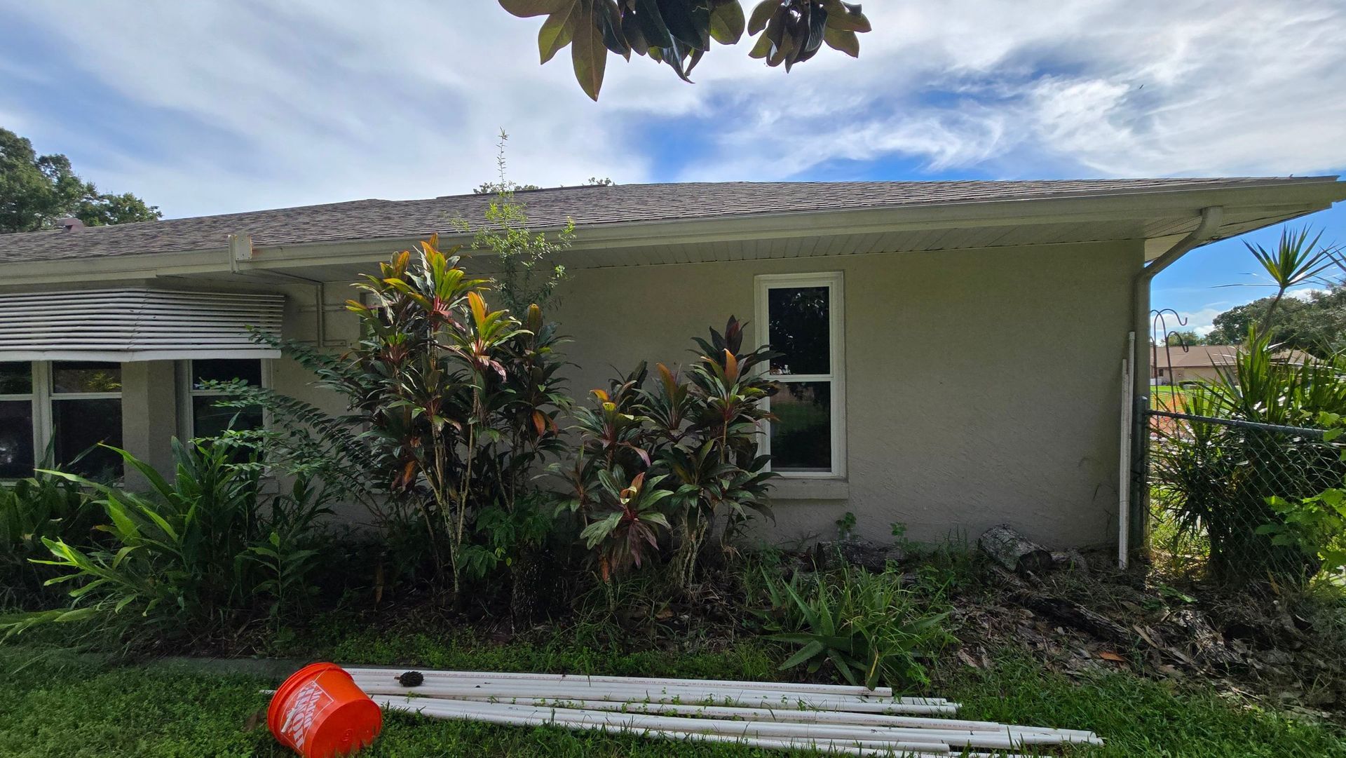 Side view of a light beige house with a cluttered roof, overgrown bushes, and a fallen bucket on the lawn.