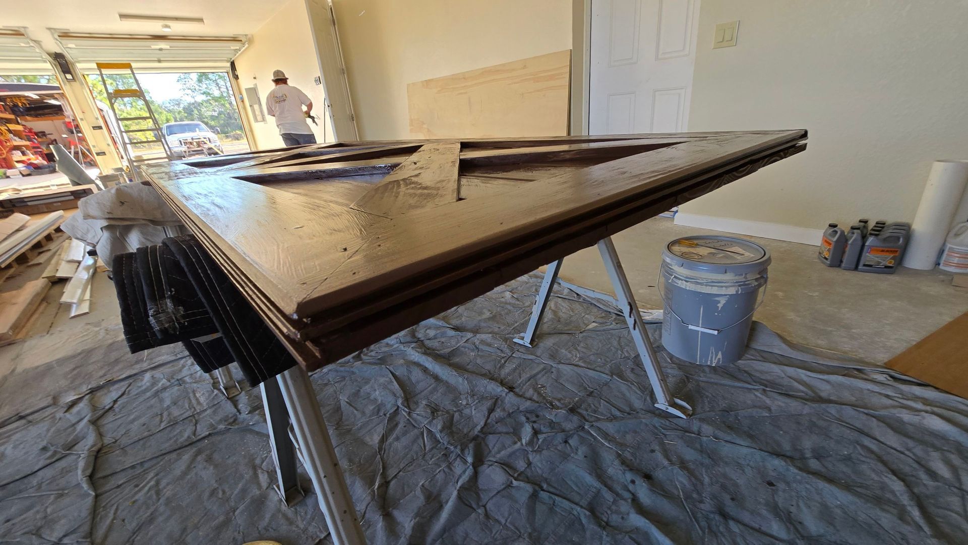 Door panels stacked on sawhorses, coated with brown finish, inside a garage. Person in background.