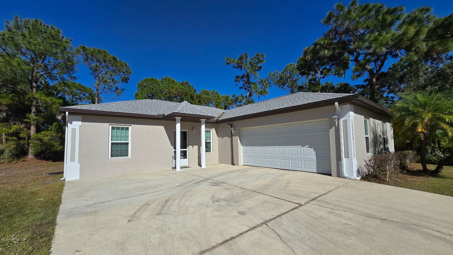 Beige house with a gray roof and a two-car garage under a blue sky, surrounded by trees.