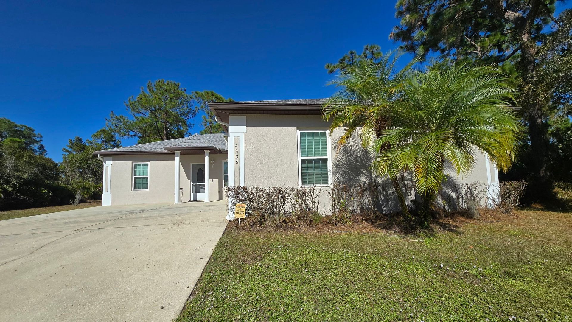 Tan house with driveway, small front yard, blue sky, and trees.