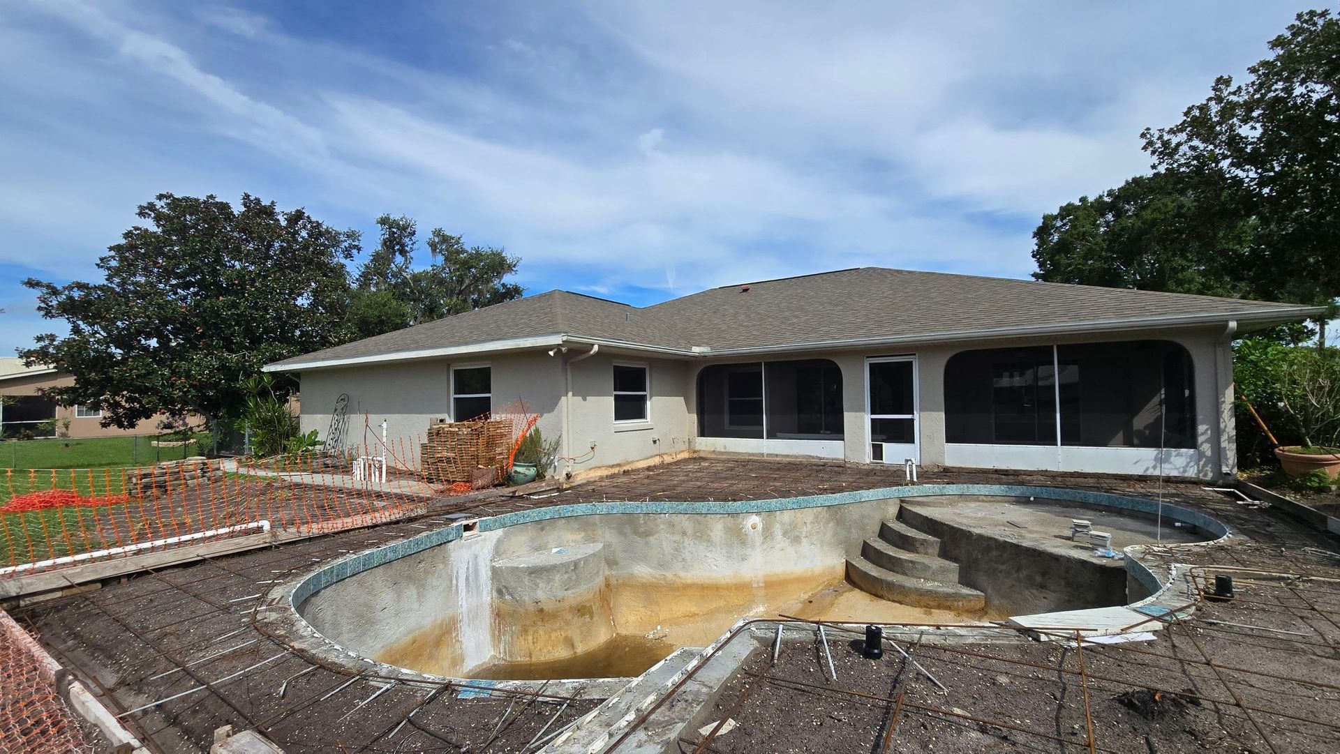 A dry, empty swimming pool in front of a house. The house has a screened-in porch and a gray roof.