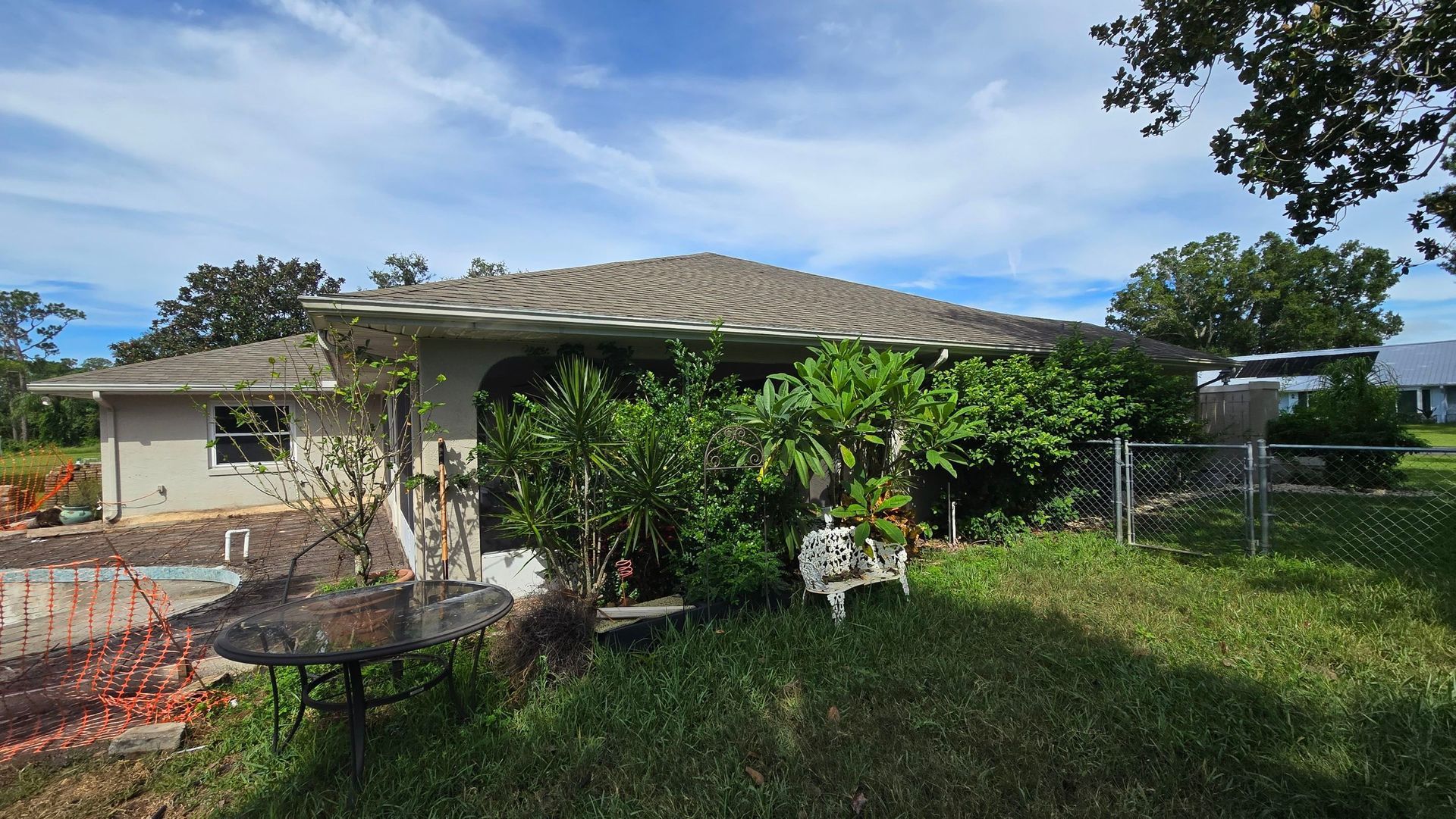 Overgrown, single-story house with a tile roof, surrounded by bushes and a chain-link fence, under a partly cloudy sky.