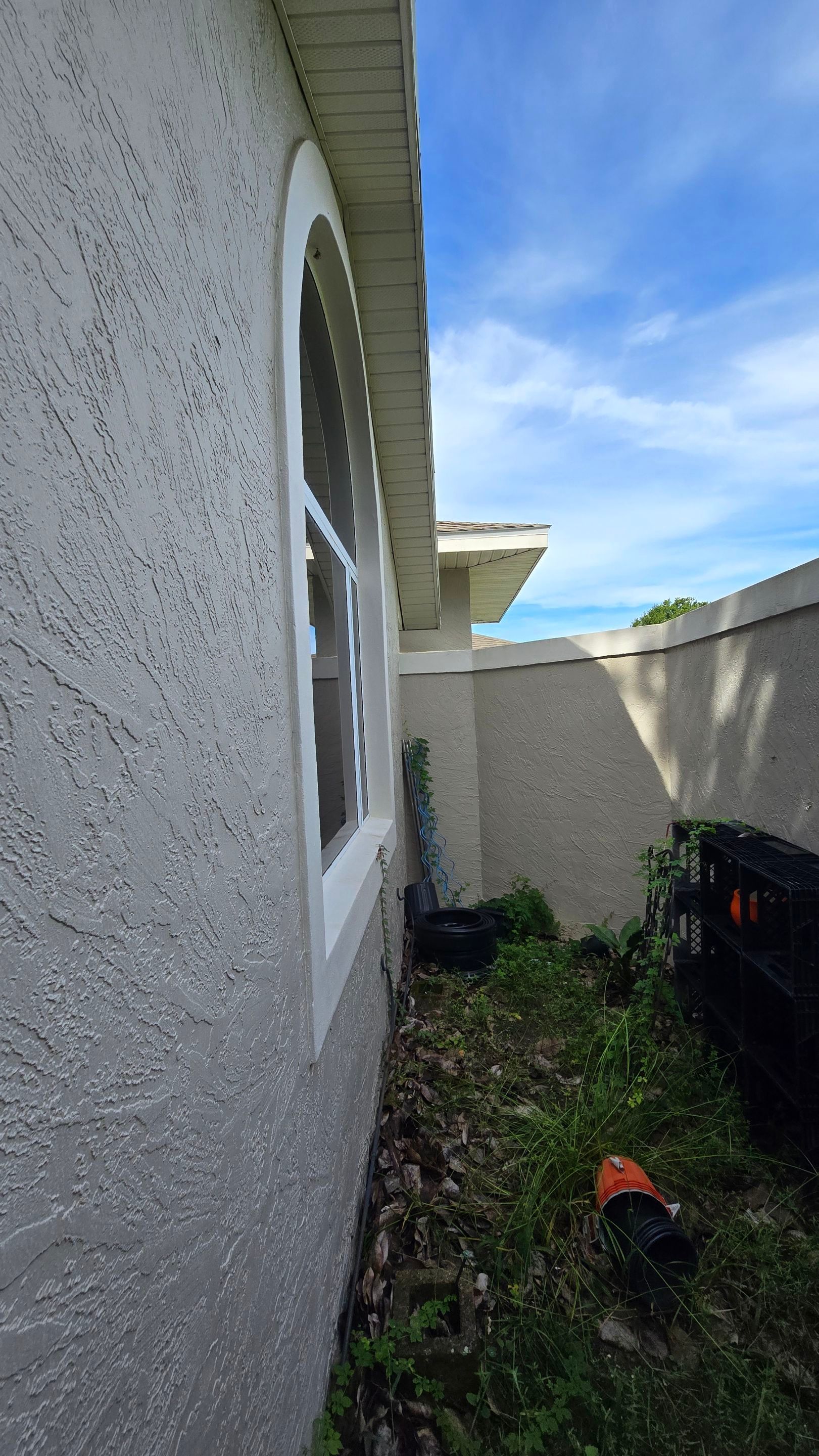 Exterior view of a stucco house with arched window, overgrown vegetation, and a clear sky.