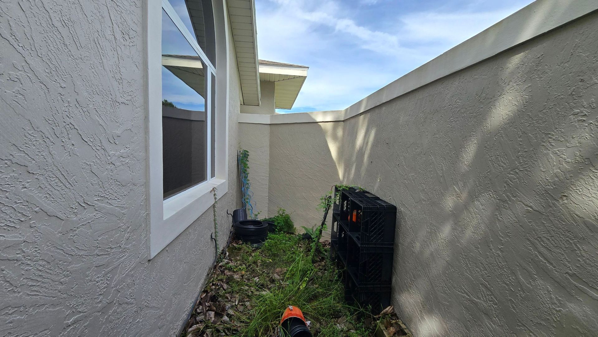 Narrow exterior space between two textured beige walls, window on left, overgrown weeds and a black shelf along the ground.