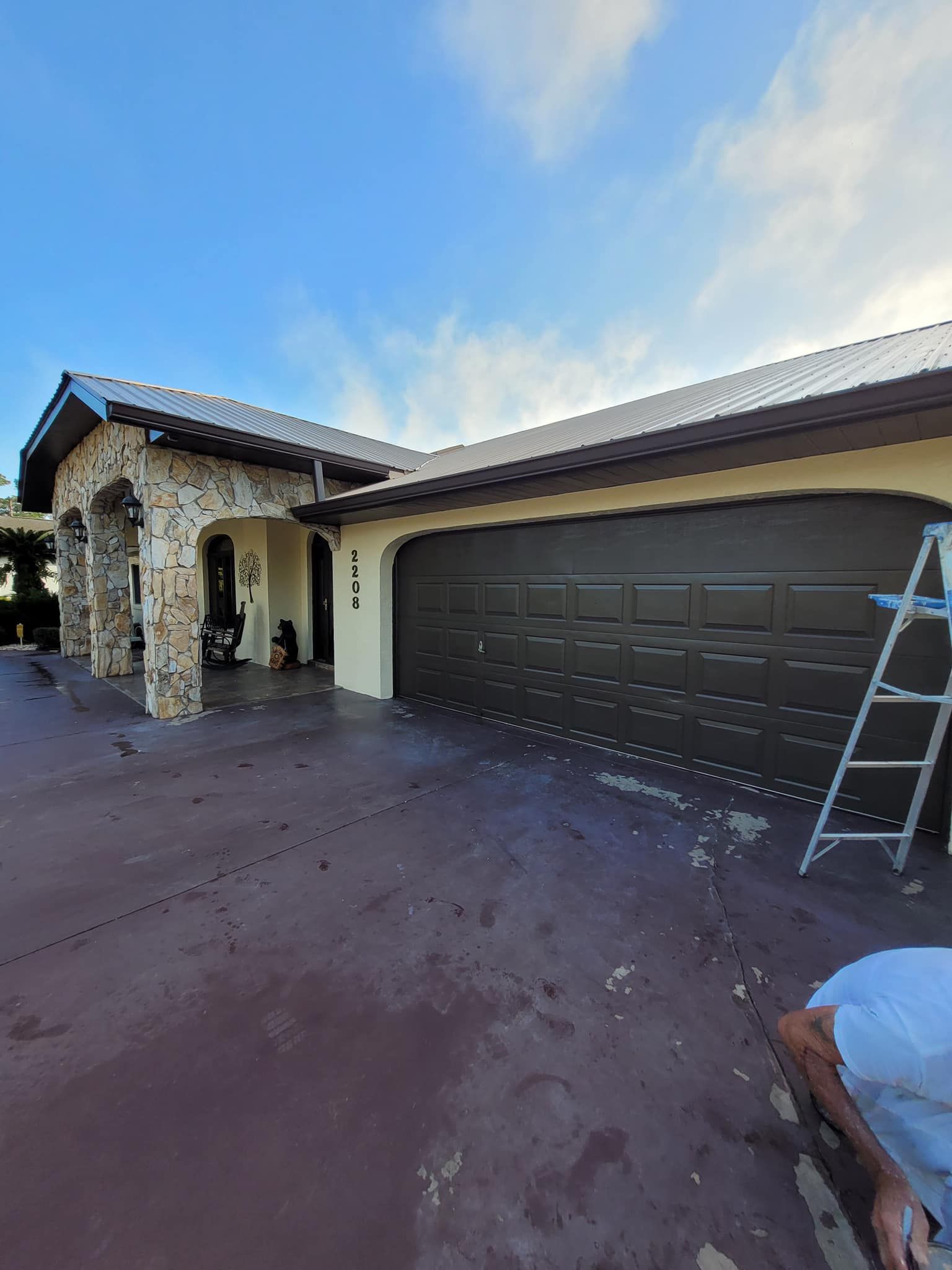 House exterior with brown garage door, arched entry, and brown driveway. A person on a ladder.