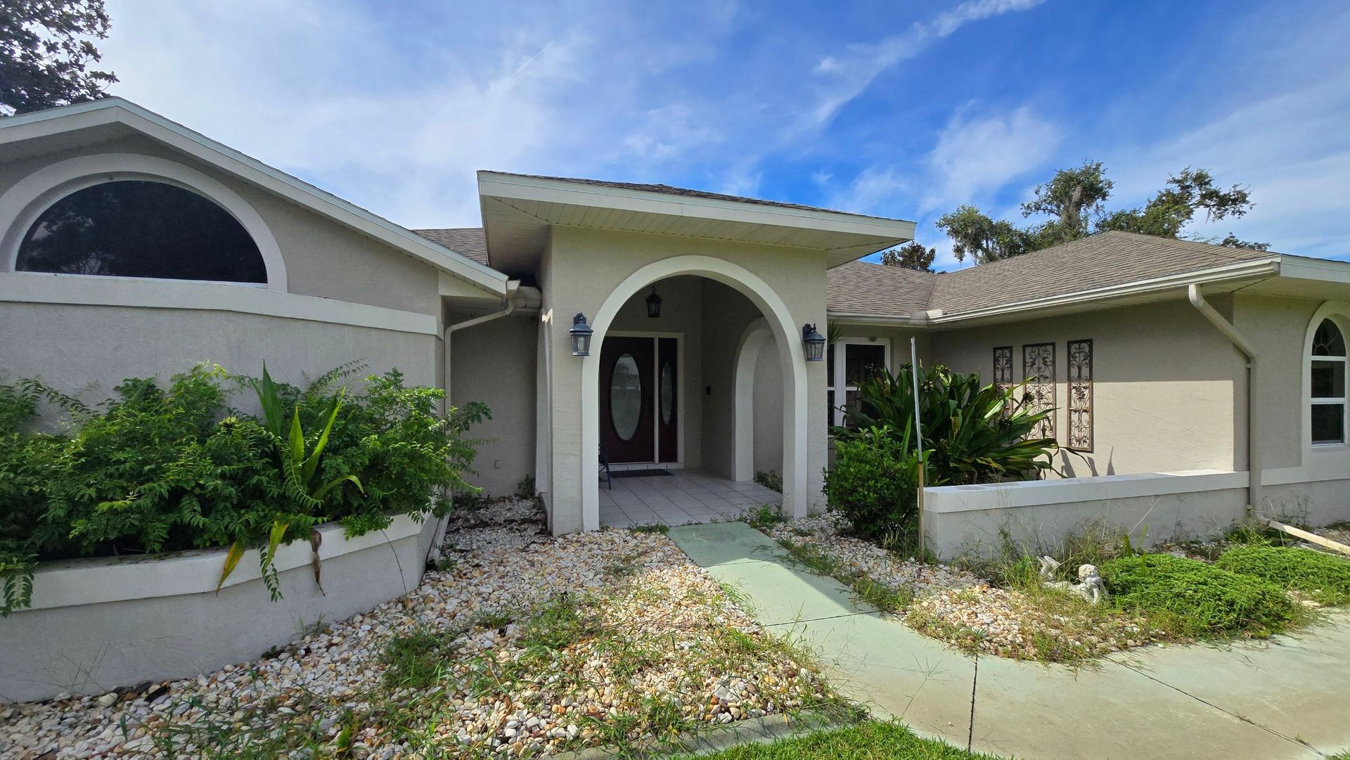 Tan stucco house with arched entry and large window, greenery, and blue sky.