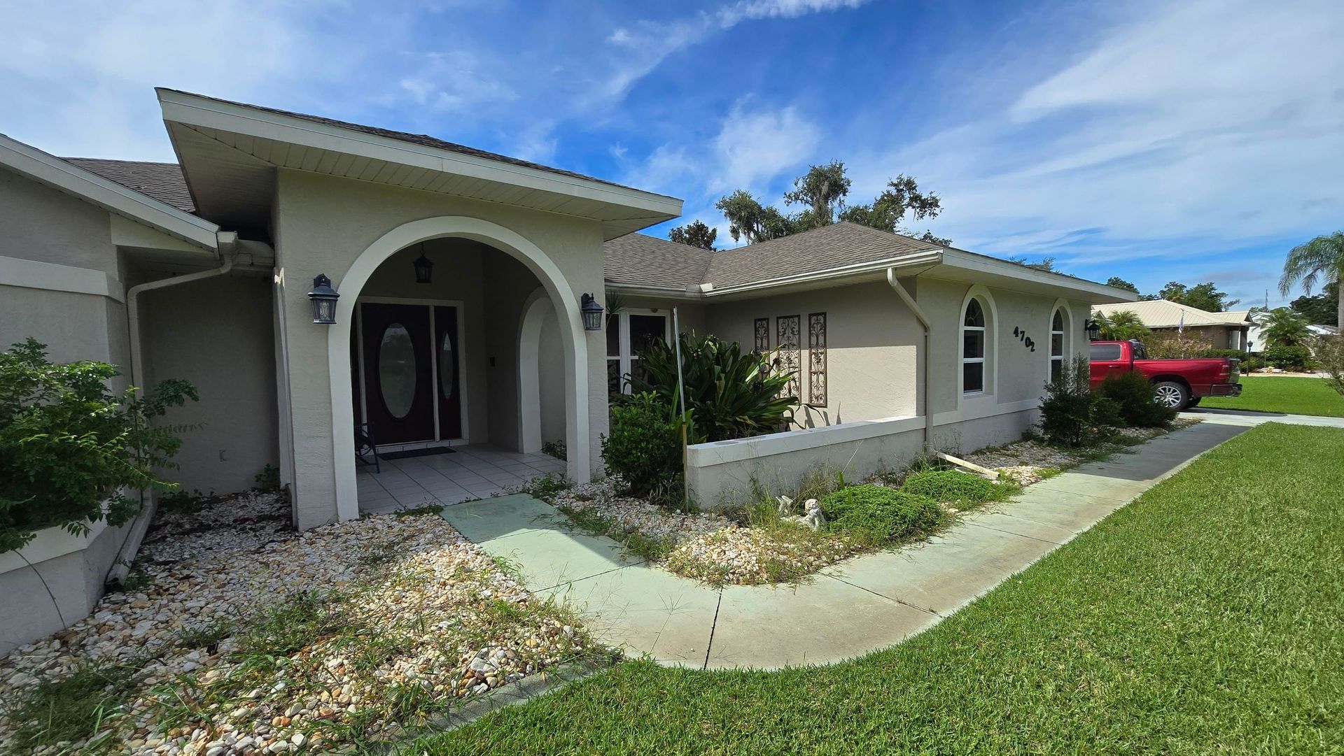 Tan ranch house with arched entryway, small yard, and red truck in the distance.