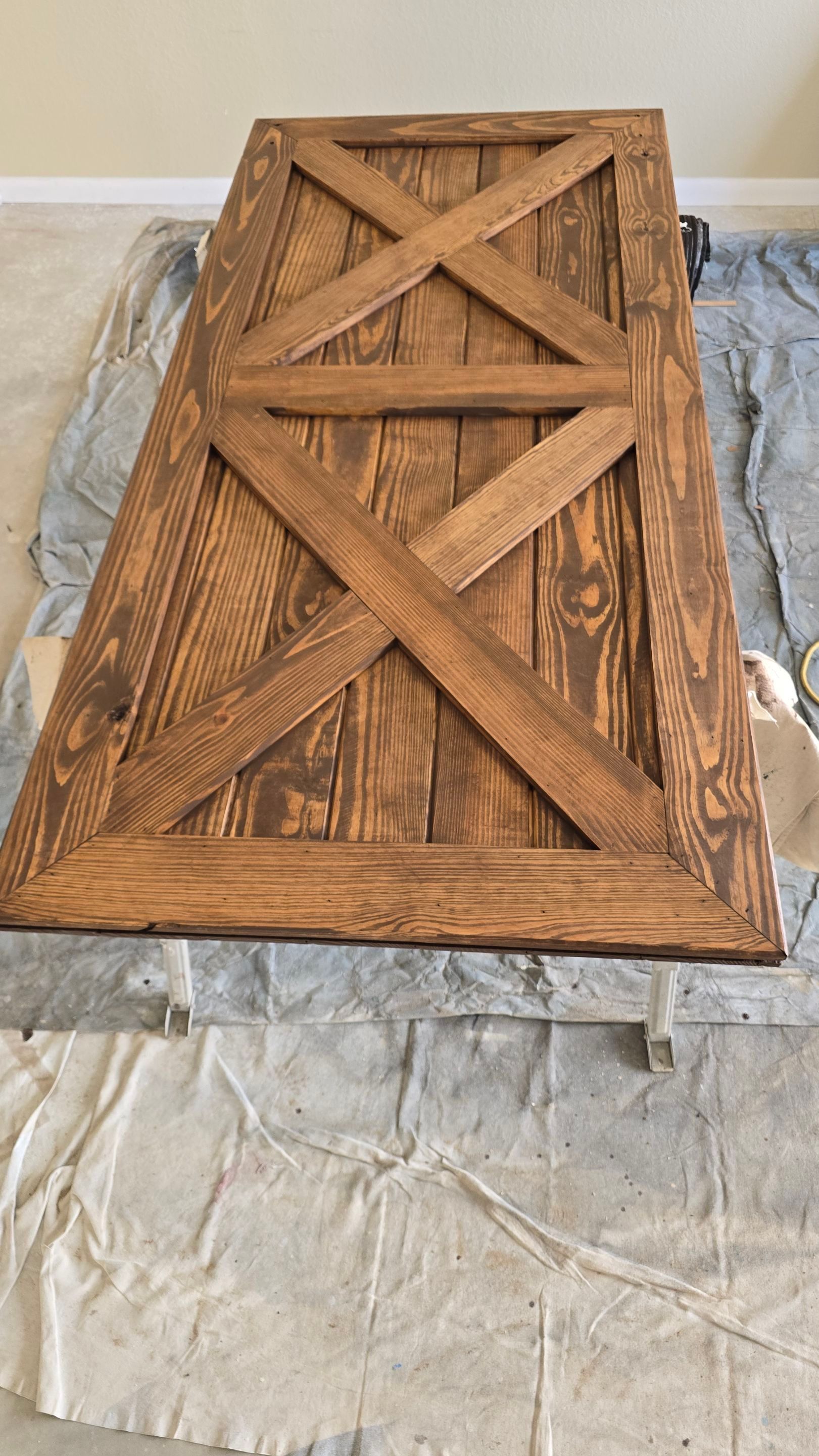 Wooden door with an X-shaped design, stained brown, lying on a drop cloth.