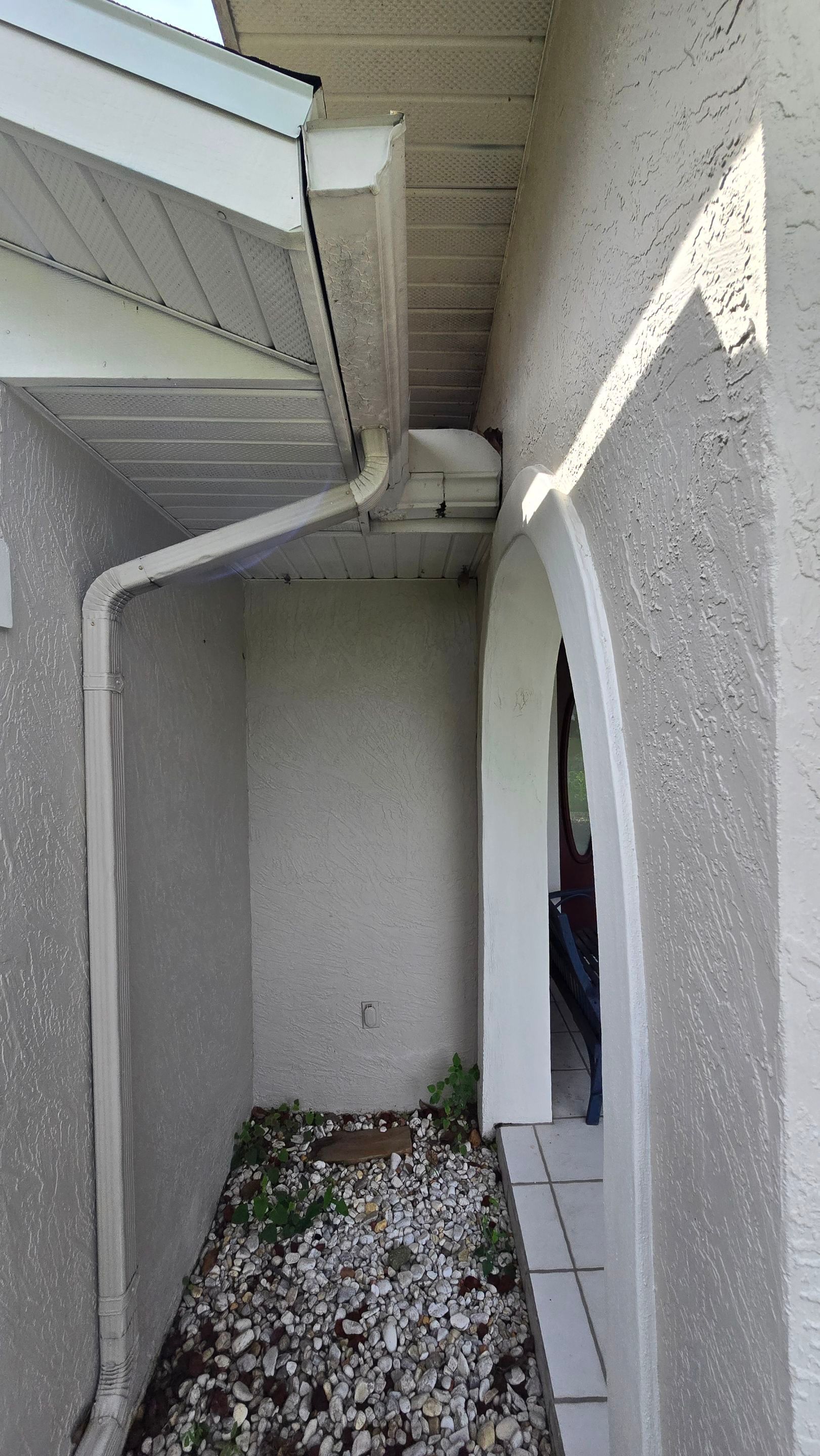 Exterior view of a house entry with a gutter, downspout, and textured stucco walls.  Gravel bed at the base.
