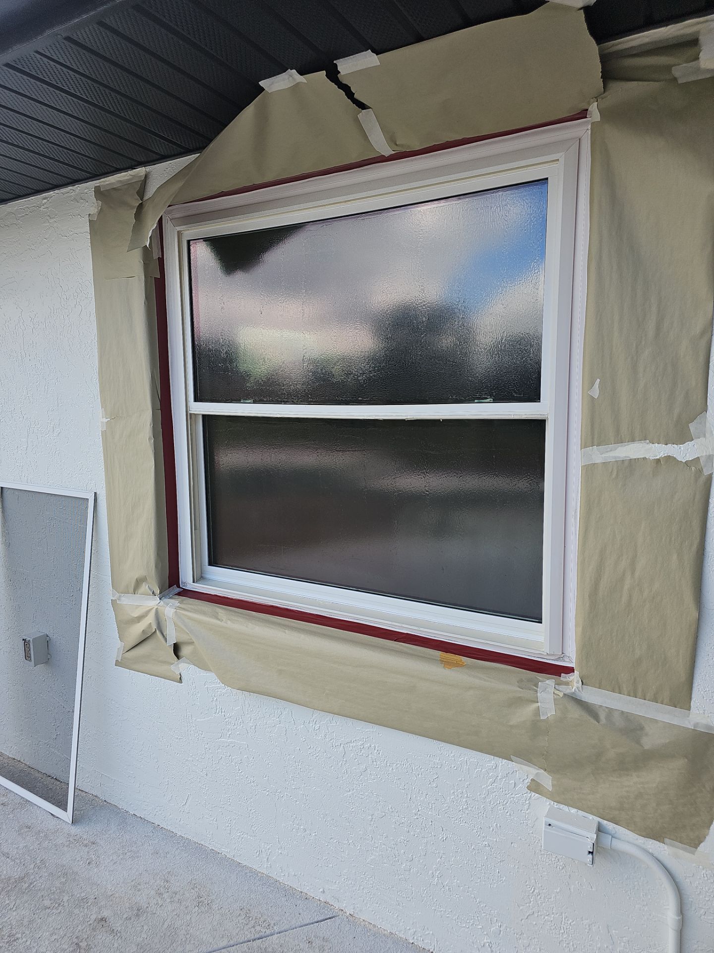 Window masked with tape and paper, ready for painting. Red trim, white frame, stucco wall.