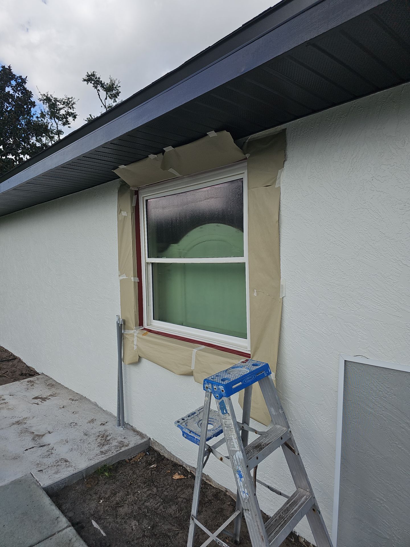 Window being painted with masking around it; a ladder stands beside.