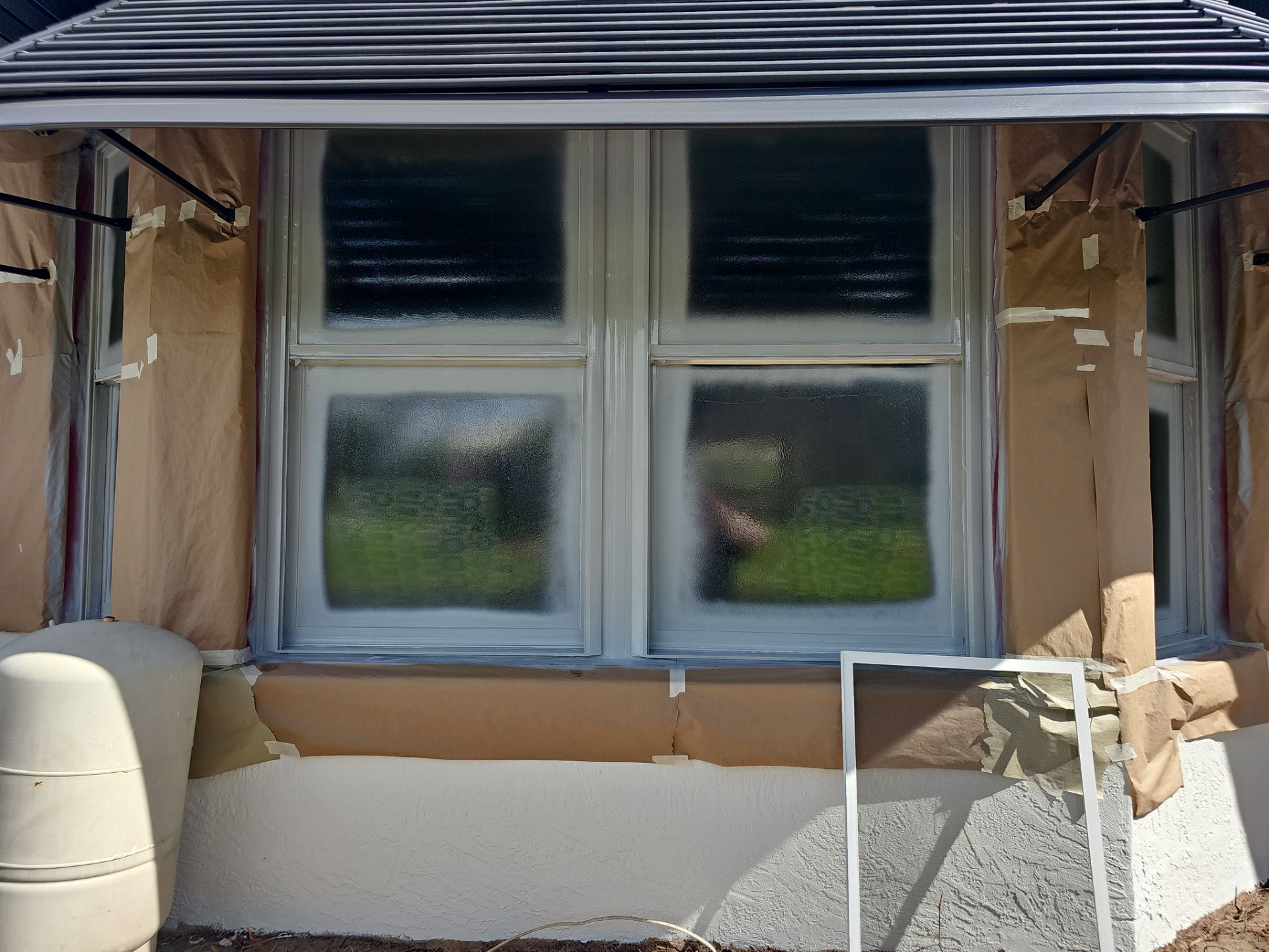 Window being painted white, masked with brown paper. Exterior view with house and foliage.