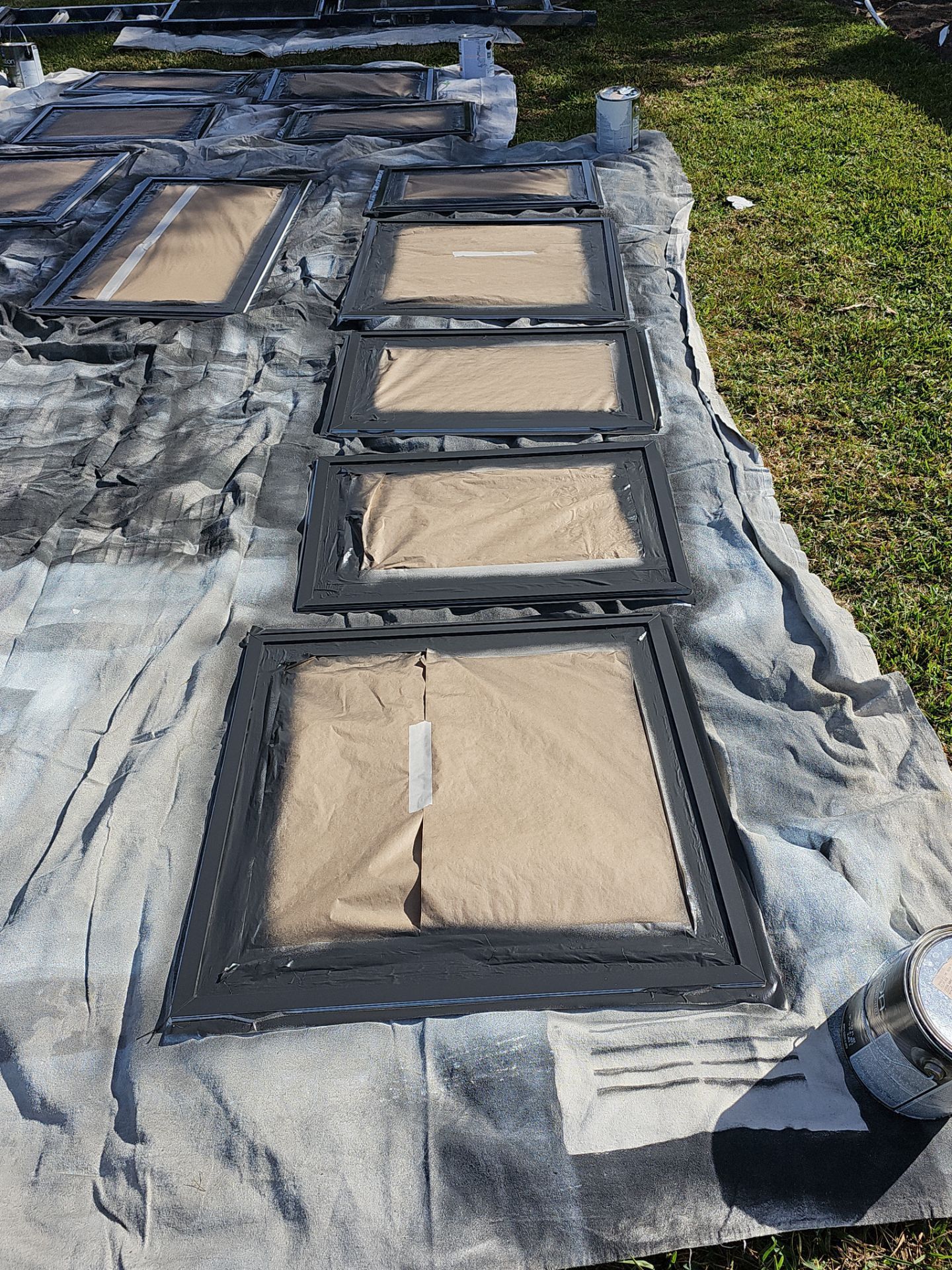 Painted cabinet doors drying on a tarp outdoors. The frames are dark gray, with brown paper covering the centers.