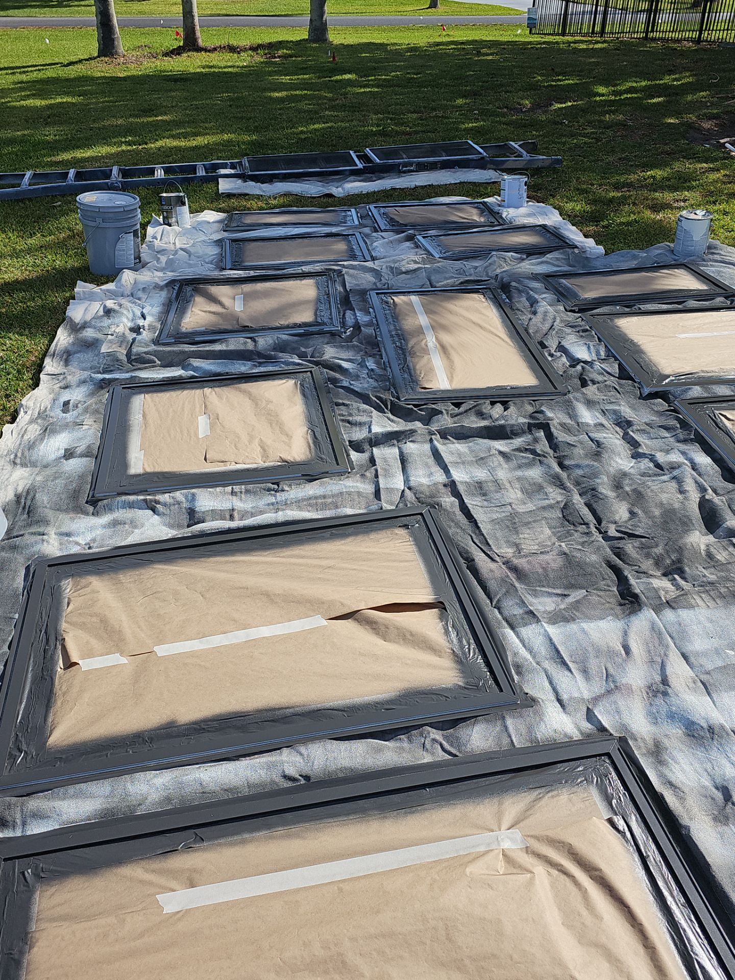 Black picture frames laid out on a tarp and being spray-painted outdoors.