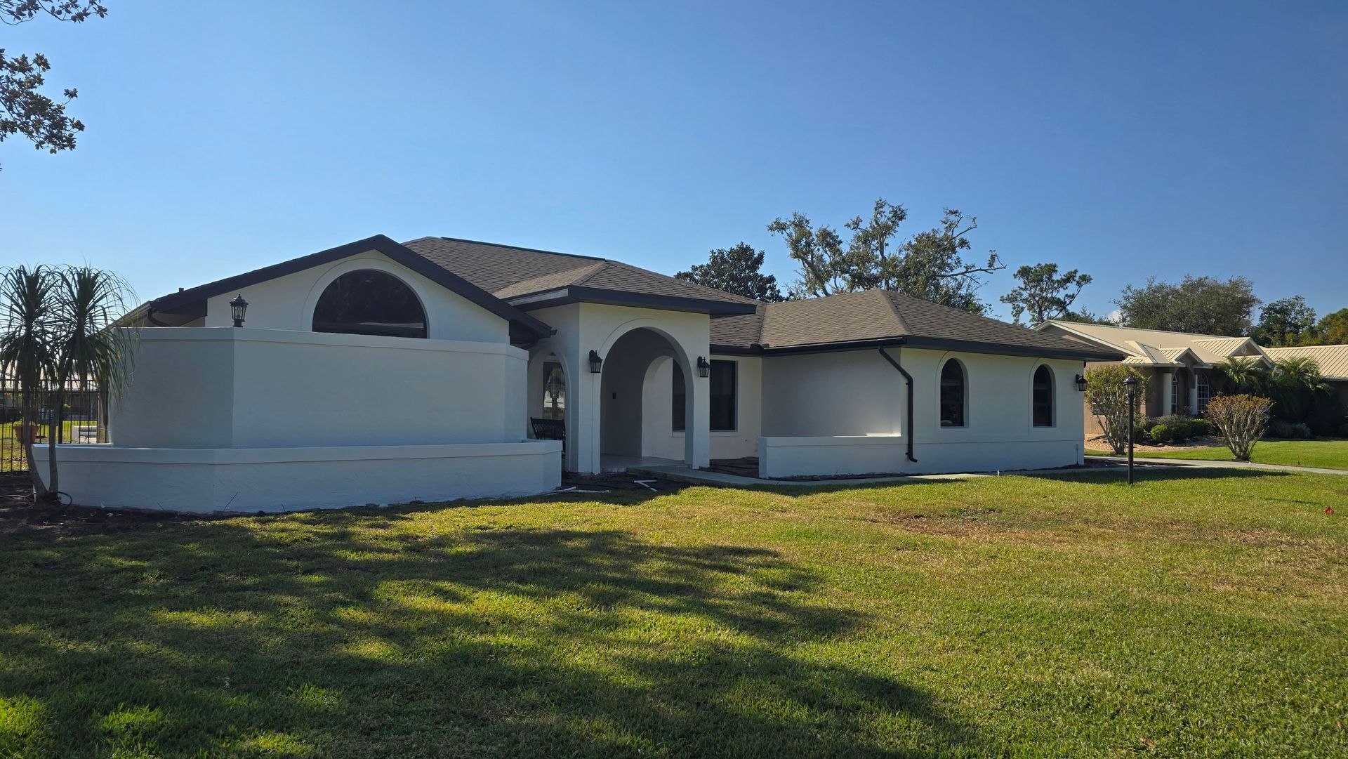 White stucco house with dark roof, arched doorway and windows, on a grassy lawn under a blue sky.