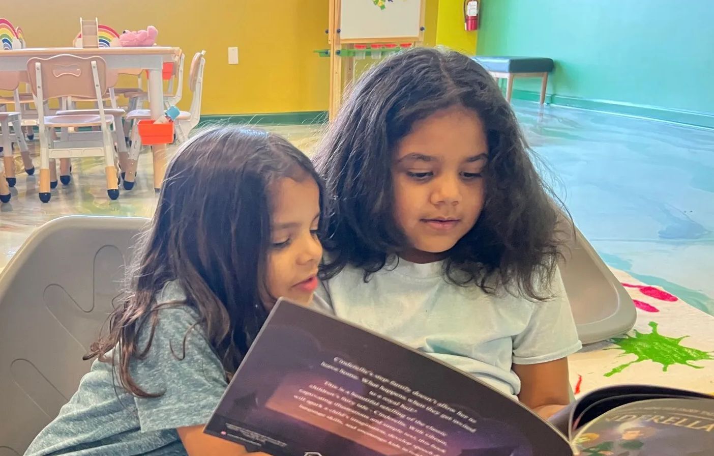 Two young girls reading a book together indoors. They are sitting and looking intently.