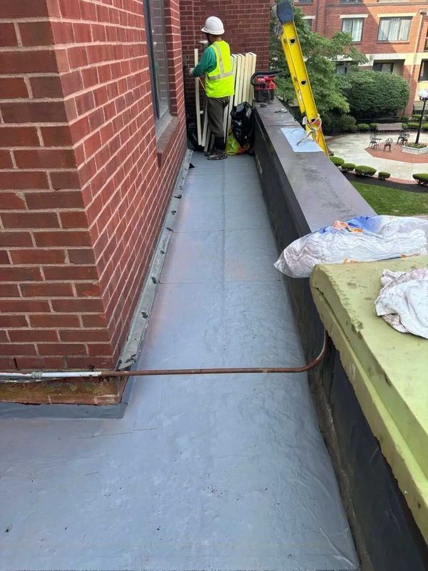 Rooftop construction: A worker in high-vis gear by a brick building. Grey flooring installed, tools, and ladder present.