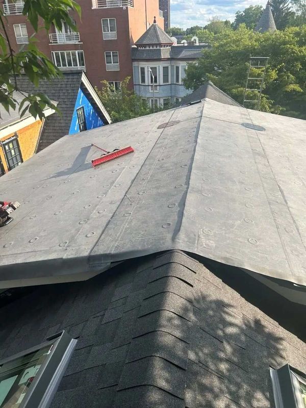 Rooftop with dark shingle and flat asphalt sections, red tool, buildings in background on a sunny day.