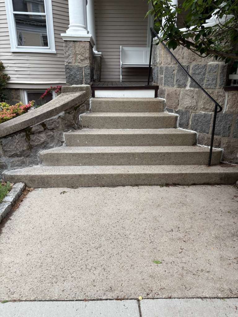 Concrete steps leading to a house entrance with stone walls and a black handrail.