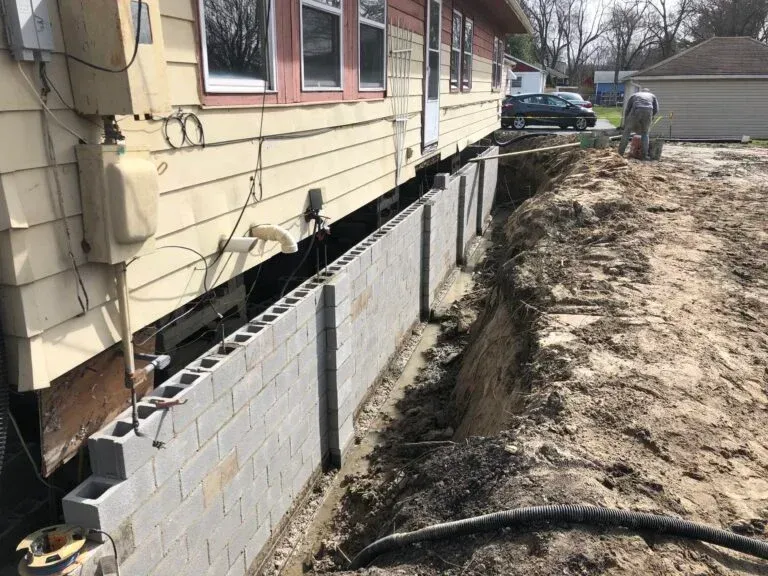 Construction site: a house raised, a cinder block retaining wall under construction. Dirt and a person visible.