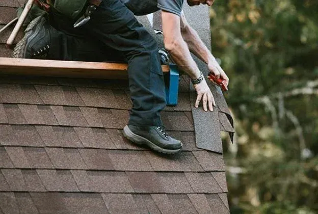 Roofer on a brown shingle roof, cutting shingles with a utility knife.