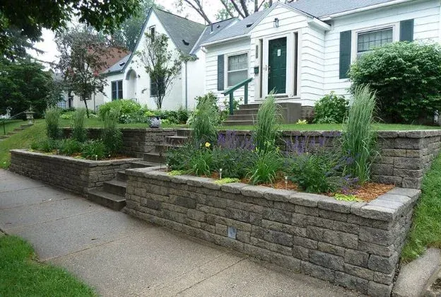 Low-lying brick retaining walls with plants in front of a white house with dark green door.