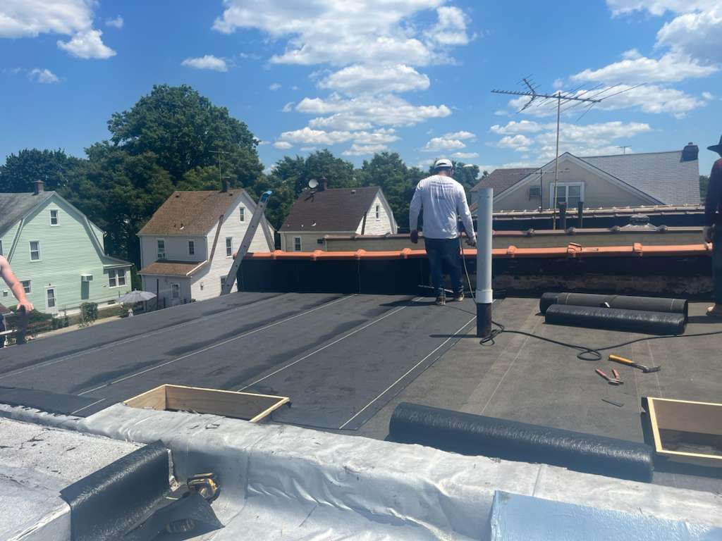 Workers on a flat roof installing roofing material under a partly cloudy sky.