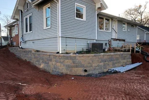 House with gray siding and a newly built retaining wall, brown soil in foreground.