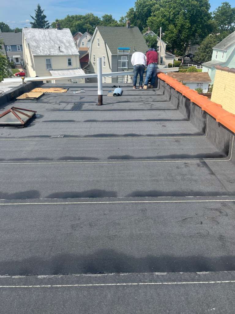 Three workers on a black flat roof, installing or repairing it. Buildings in the background.