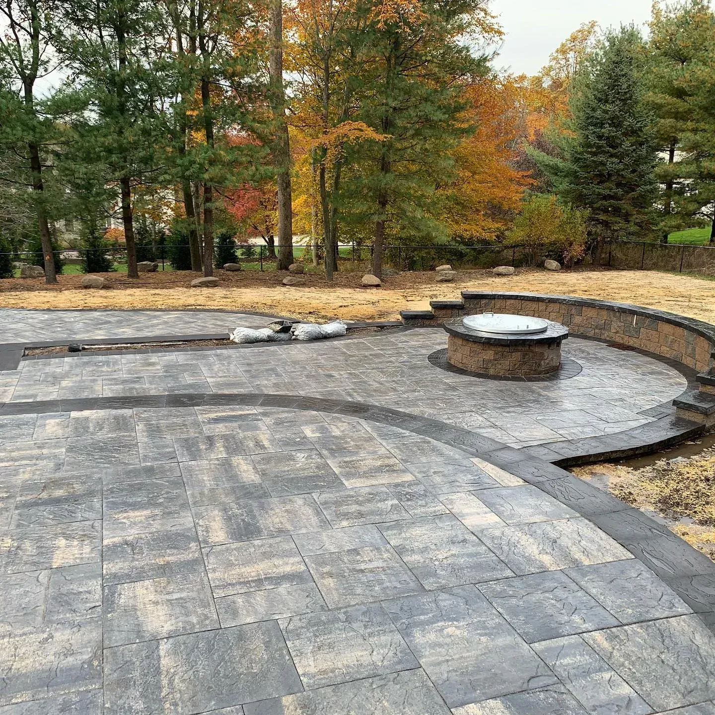 Stone patio with a curved wall and fire pit, surrounded by trees with autumn foliage.