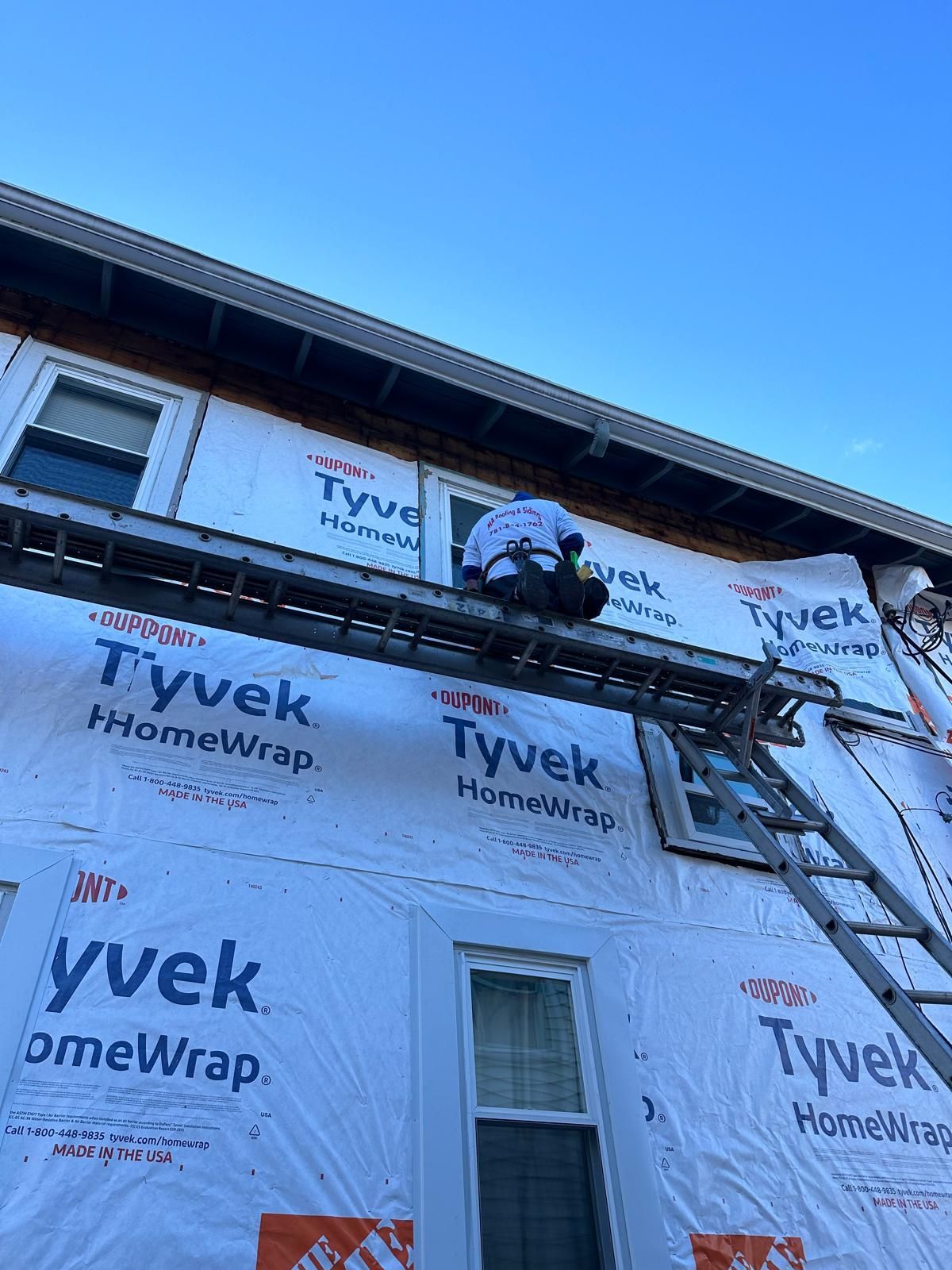 Person on a scaffold working on a house exterior; Tyvek wrap visible. Blue sky.