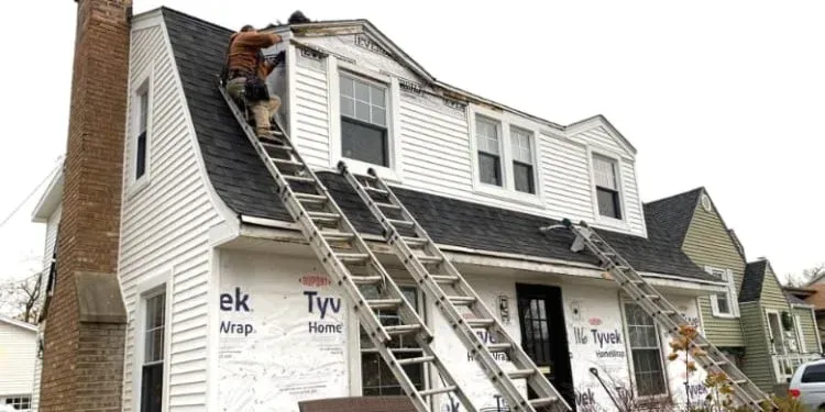 Workers on ladders repairing a house roof, with white siding and a brick chimney.