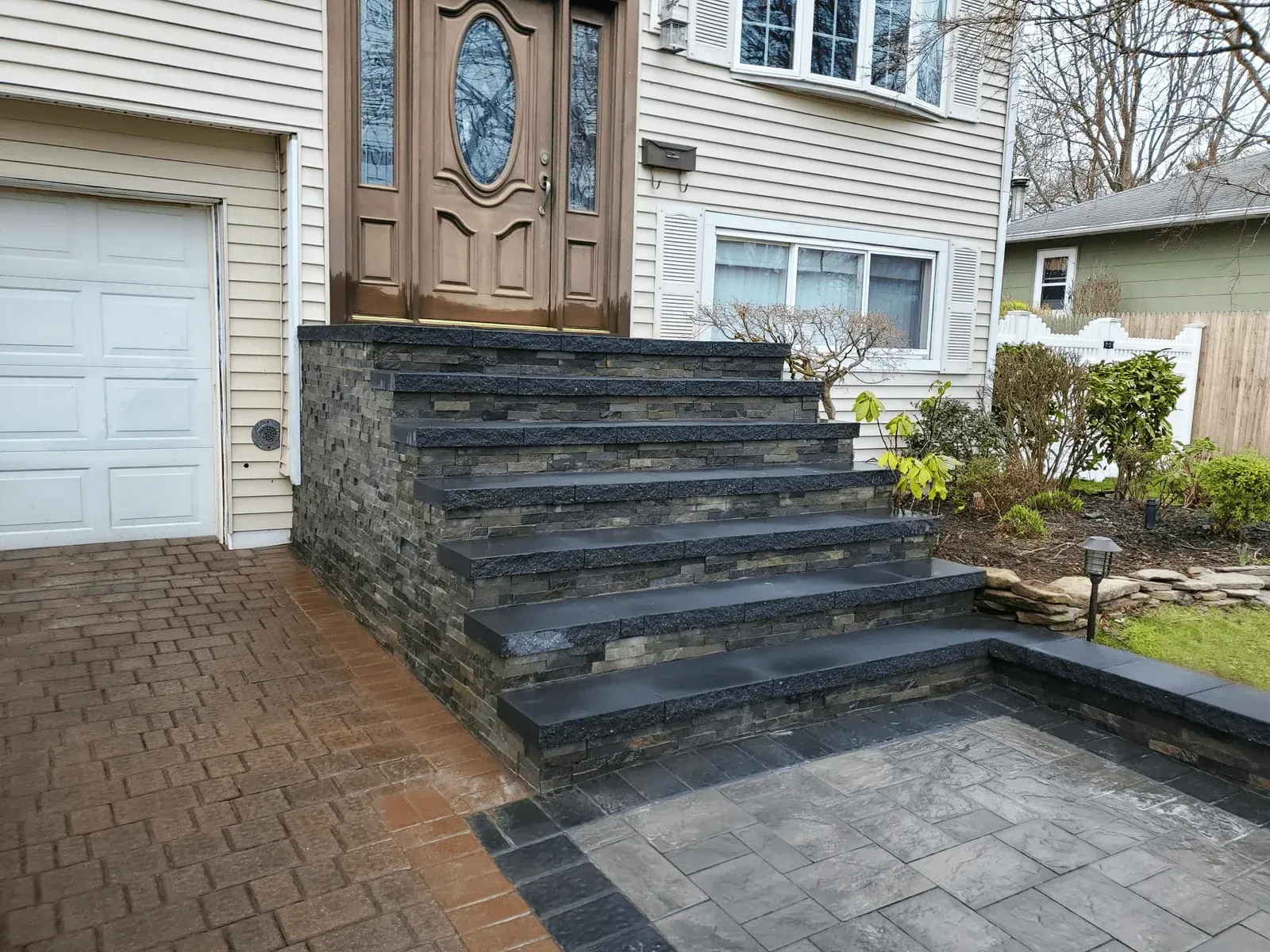 Stone steps leading to a brown front door of a light-colored house. Brick driveway and grass lawn.